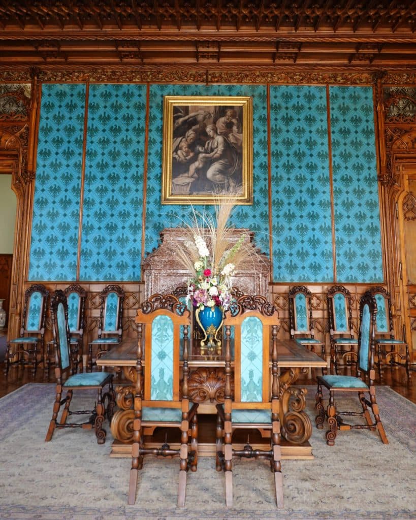 Een sierlijke eetkamer met ingewikkeld gesneden houten stoelen en tafel, allemaal met groenblauwe bekleding - geschikt voor prinses kleding. De groenblauwe muren met een zwart patroon, houten lambrisering en bloemstukken roepen de elegantie op van de grootse interieurs van Tsjechië.