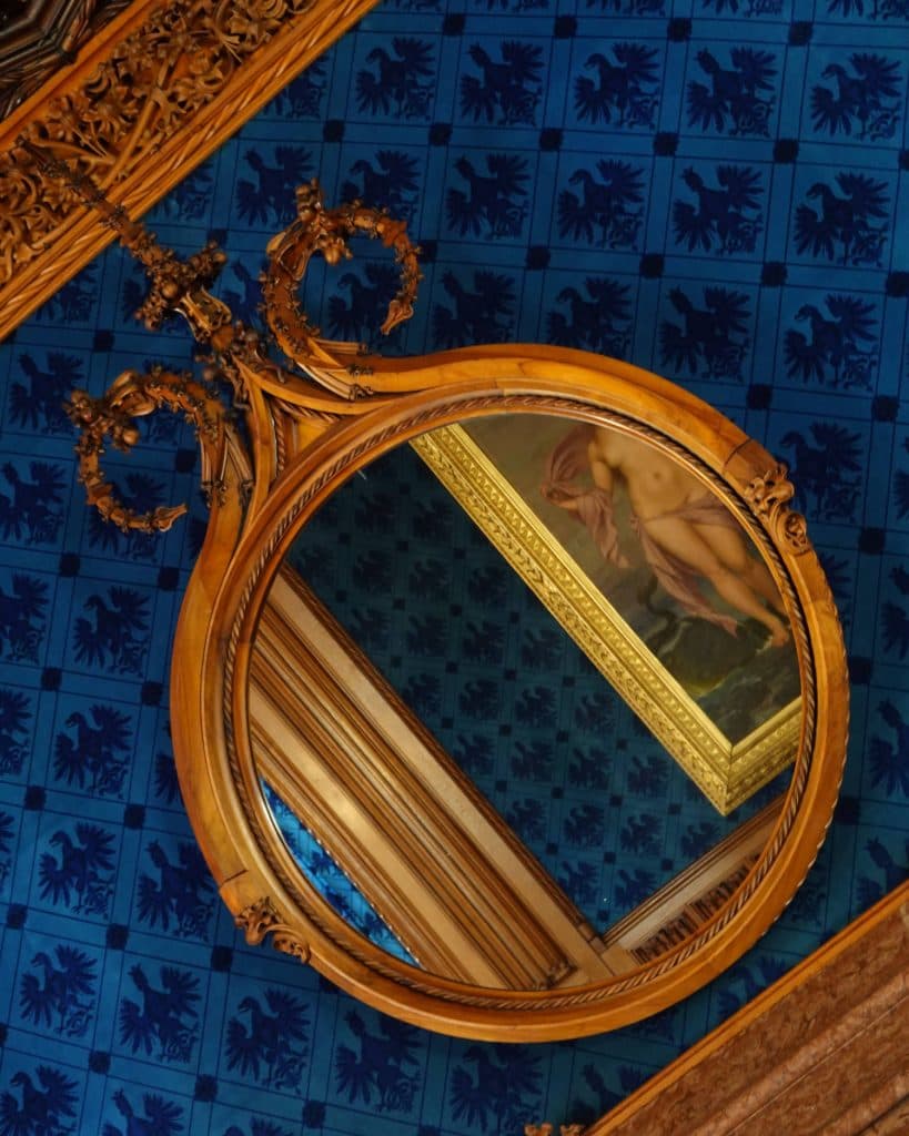Een ronde, sierlijke houten spiegel hangt aan een rijkelijk versierd plafond met blauw behang met patroon. De spiegel weerspiegelt een deel van een klassiek schilderij van een prinses en een goudomrande lijst, wat een artistiek tintje toevoegt aan het elegante tafereel.