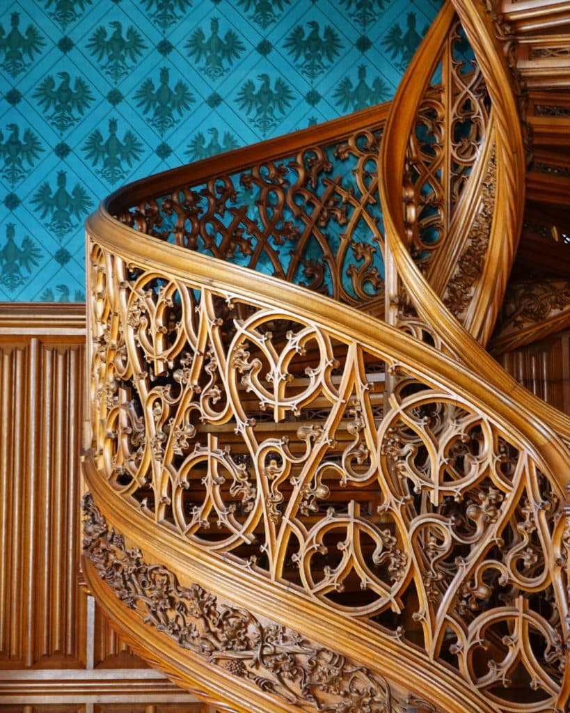 Een close-up van een sierlijke houten wenteltrap met ingewikkeld houtsnijwerk tegen muren versierd met blauw behang met heraldische adelaarsmotieven die doen denken aan kastelen in Tsjechië en houten lambrisering eronder.