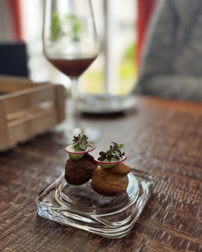 Twee kleine, elegant opgediende hapjes met schijfjes radijs en microgroenten zitten op een doorzichtige glazen schaal. Op de onscherpe achtergrond roepen een houten kist, een rood wijnglas en een diner in een zachte trui gezellige avonden in Tsjechië op.