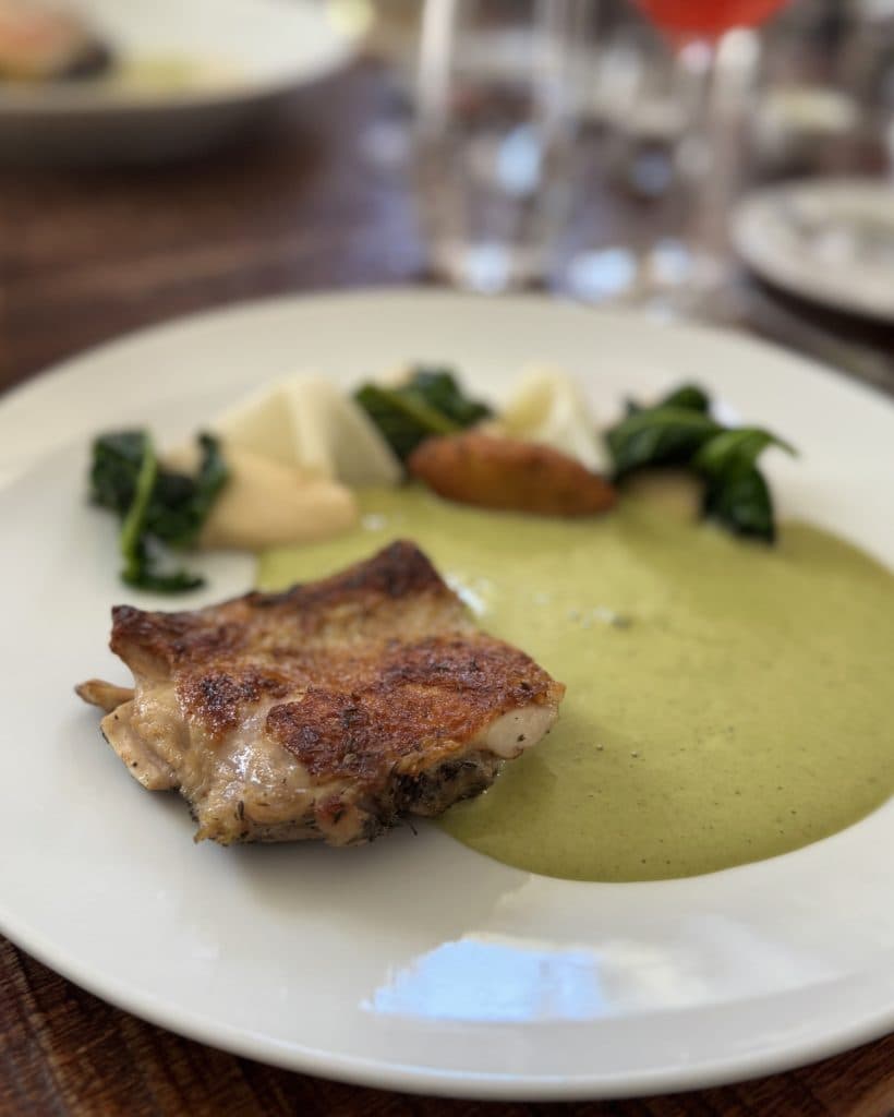 Op een wit bord ligt een stuk geroosterde kip met knapperige huid, geserveerd met groene saus, gesauteerde groenten, geroosterde aardappel en een paar stukjes lichtgekleurde wortelgroenten.