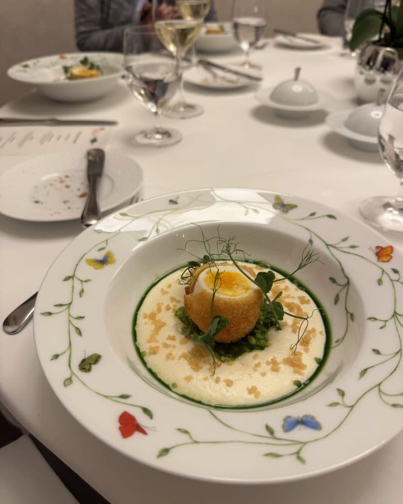 Een gastronomisch gerecht is opgediend in een bloemige porseleinen schaal: een gouden bol op groene erwten en romige witte saus, gegarneerd met microgroenten. De tafel is elegant gedekt als voor een prinses, met wijn, waterglazen, bestek en een ander soortgelijk gerecht op de achtergrond.