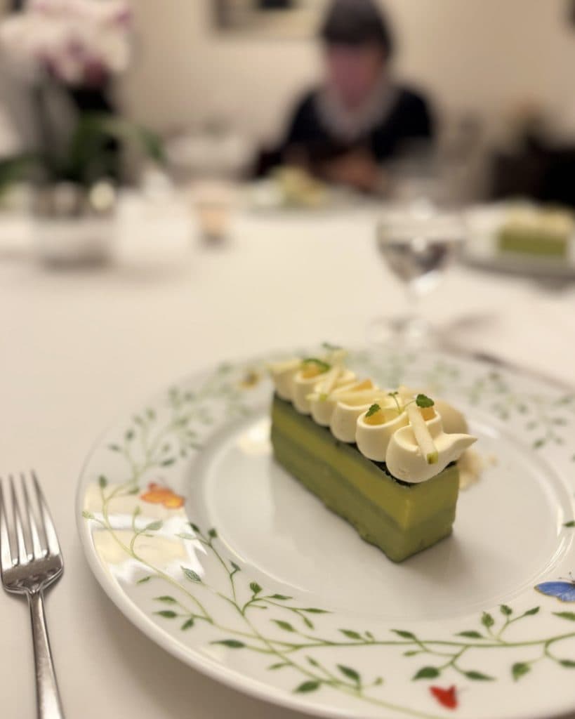Op een bord met bloemmotief ligt een rechthoekig pistachenootjesdessert met slagroom en kleine groene garnering. Een vork is links geplaatst. Op de onscherpe achtergrond dineert iemand in een prinses jurk aan een tafel met een wit tafelkleed en een glas water.