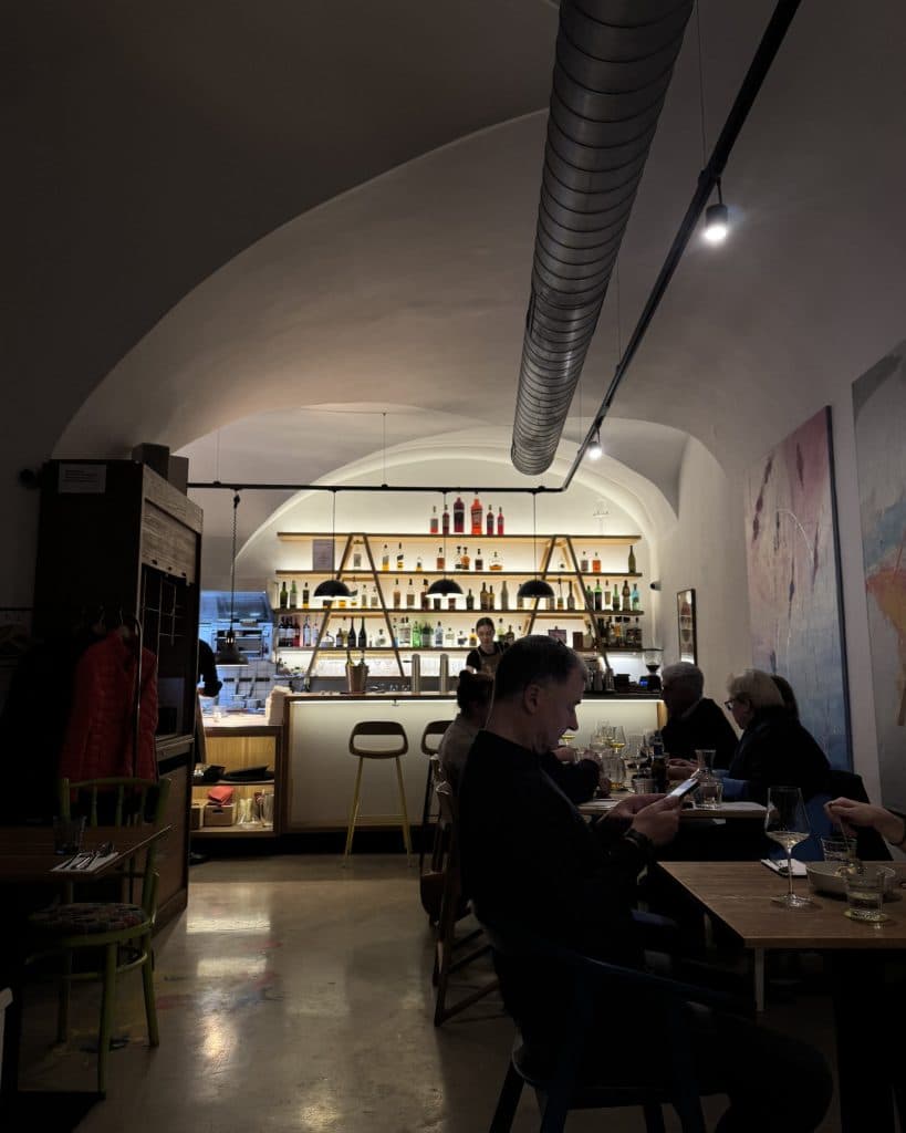 Een schemerig verlichte moderne bar met gewelfde plafonds. Planken tonen flessen en glazen achter de toonbank.