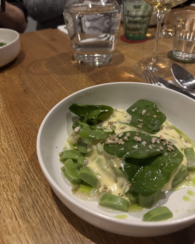 Een witte schaal op een houten tafel bevat een salade met verse spinaziebladeren, sperziebonen, romige gele dressing en bestrooide zonnebloempitten - perfect voor een voordelige vakantie geïnspireerd door Tsjechië. Op de achtergrond zie je een glas water en een wijnglas.