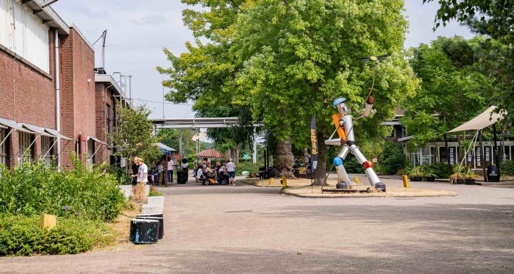 Een levendige binnenplaats met een hoog, kleurrijk robotbeeld dat een dirigeerstokje vasthoudt, laat de invloed van de automatische concepten zien. Mensen verzamelen zich links bij een café in de schaduw en genieten van de levendige sfeer. Bomen en groen omringen het tafereel, met bakstenen en glazen gebouwen op de achtergrond onder een stralende hemel.
