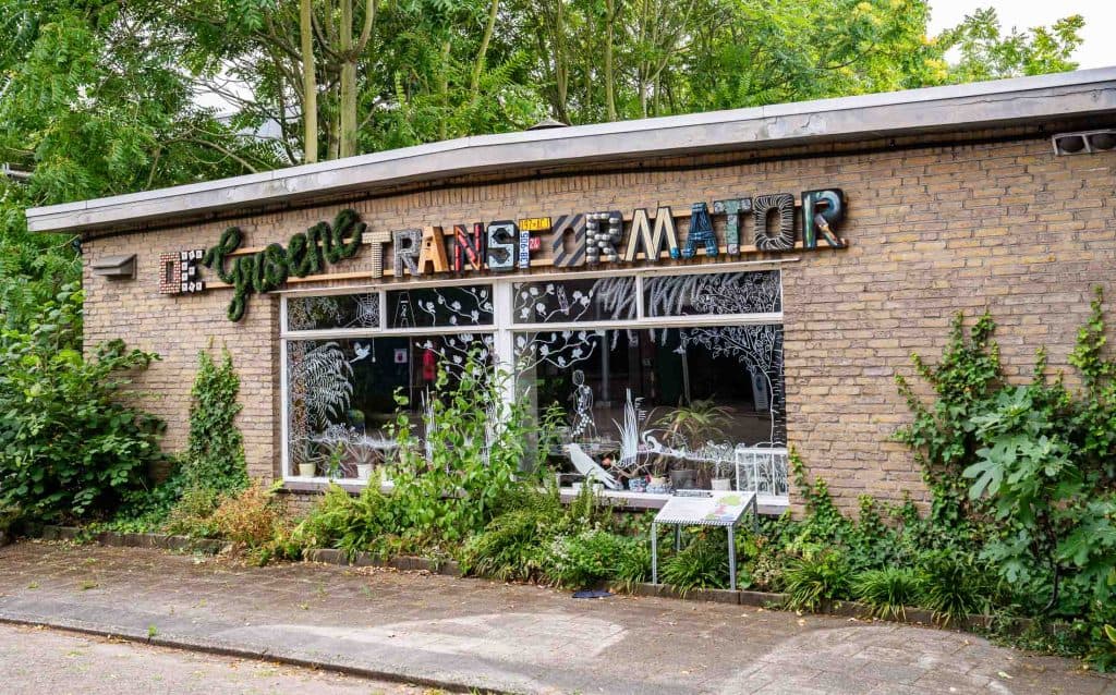 Een bakstenen gebouw met het opschrift „Cyberspace Transformatior“ in kleurrijke letters van verschillende stijlen boven grote ramen valt op in de straat. De ramen zijn versierd met gedetailleerde witte patronen, die verwijzen naar de aanwezigheid van automatische concepten binnenin. Rondom het gebouw groeien planten en groen, en de straat ervoor is leeg.