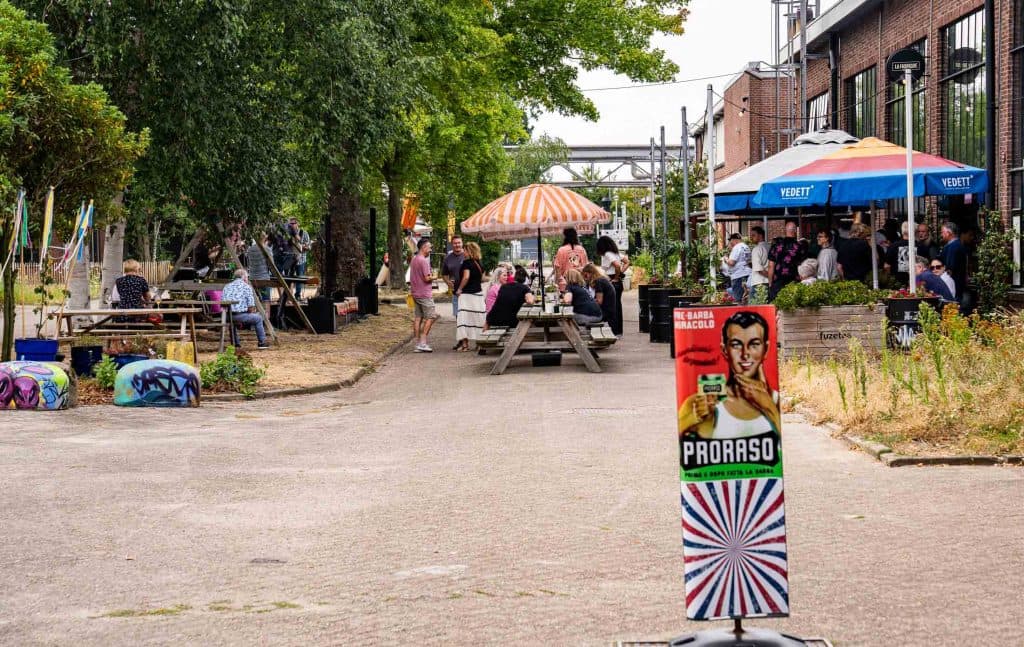 Een buitenopname met mensen die aan picknicktafels onder parasols zitten bij een bakstenen gebouw, waar automatische oplossingen voor zitplaatsen en schaduw een comfortabele omgeving creëren. Het terrein wordt omzoomd door bomen. Op de voorgrond staat een kleurrijk „Proraso“-bord. Sommige mensen zijn aan het eten, aan het kletsen of aan het ontspannen, wat bijdraagt aan de levendige, ongedwongen sfeer.