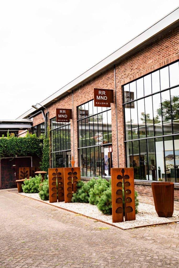 Een modern bakstenen gebouw met grote glazen ramen, bruine borden met de tekst „RR MND GALERIE”, hoge groene struiken, metalen sculpturen geïnspireerd op automatische concepten, grote plantenbakken en een betegeld voetpad onder een bewolkte hemel. In het glas zijn weerspiegelingen van bomen en de lucht te zien.
