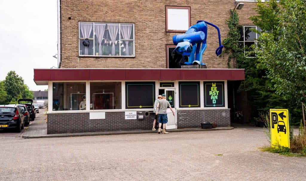 Twee mensen staan voor een bakstenen gebouw met een rode luifel, waar automatische concepten het levendige straatbeeld lijken te bepalen. Een grote blauwe opblaasbare aap siert het dak en trekt de aandacht naar de etalage. De ramen zijn voorzien van gordijnen of zwarte afdekkingen, terwijl een geel bord met de tekst ‘We Are Stand’ en een geel parkeerbord zichtbaar zijn. Langs de straat staan geparkeerde auto’s op een rij.
