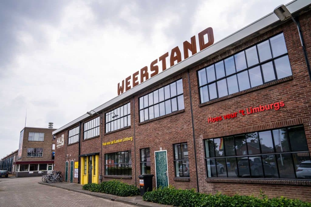 Een bakstenen gebouw in industriële stijl met grote ramen en het woord „WEERSTAND” op het dak. Gele en rode deuren, buiten geparkeerde fietsen en Nederlandse tekst op de gevel wijzen op de levendige sfeer. De lucht is bewolkt en op de achtergrond staat nog een soortgelijk gebouw – samen vormen deze plekken wellicht knooppunten voor innovatieve ideeën en automatische concepten die bepalend zijn voor modern stedenbouwkundig ontwerp.