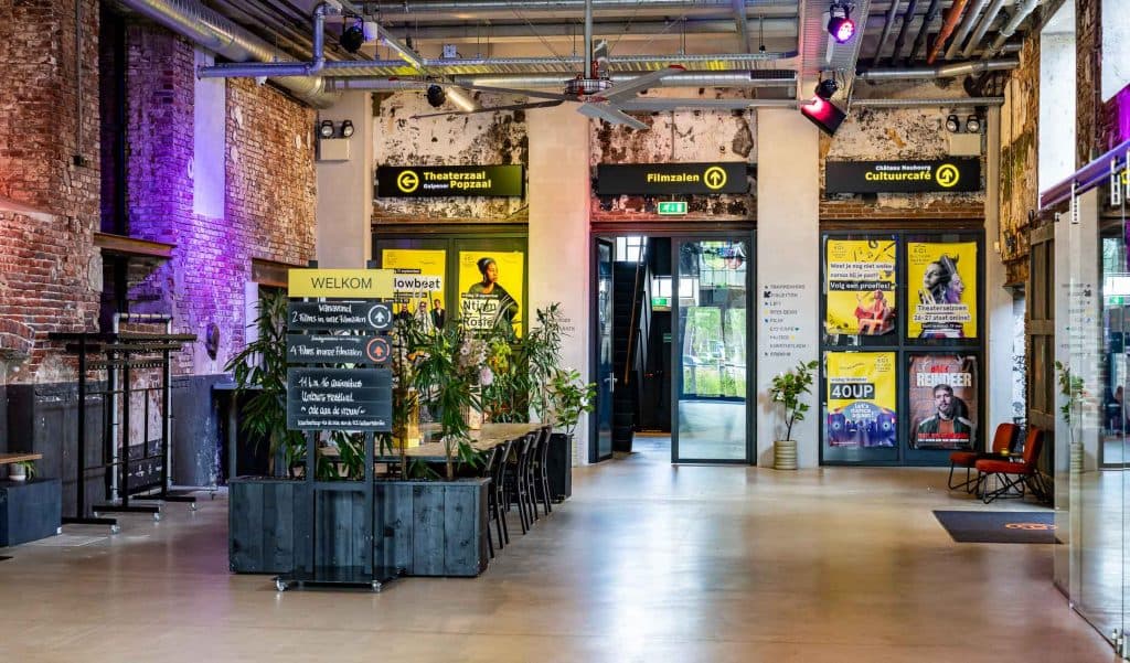 Een moderne lobby met een industriële inrichting, zichtbare bakstenen, kleurrijke lampen, planten en posters aan de muren. Borden wijzen de weg naar theaters, een bioscoop en een cultuurcafé met innovatieve automatische concepten. Grote ramen laten bij de ingang natuurlijk licht binnen.