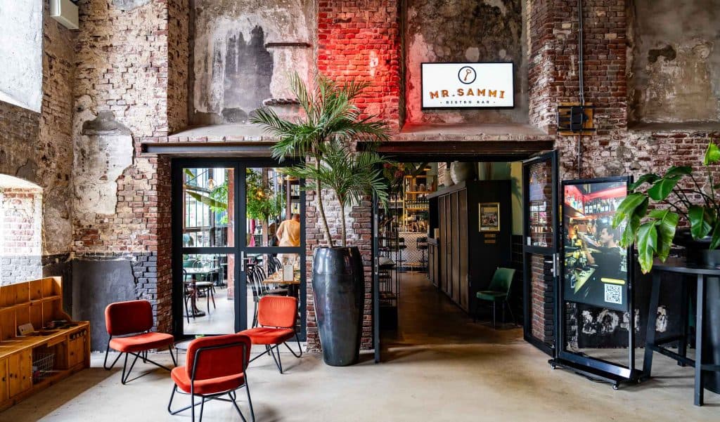 Een café in industriële stijl met bakstenen muren, hoge planten en rode stoelen. Grote glazen deuren geven toegang tot de bar binnen, waar automatische concepten naadloos in het ontwerp zijn geïntegreerd. Boven de ingang hangt een bord met de tekst „Mr. Sammi”. Warme verlichting, groen en een rustieke inrichting zorgen voor een gezellige, uitnodigende sfeer.