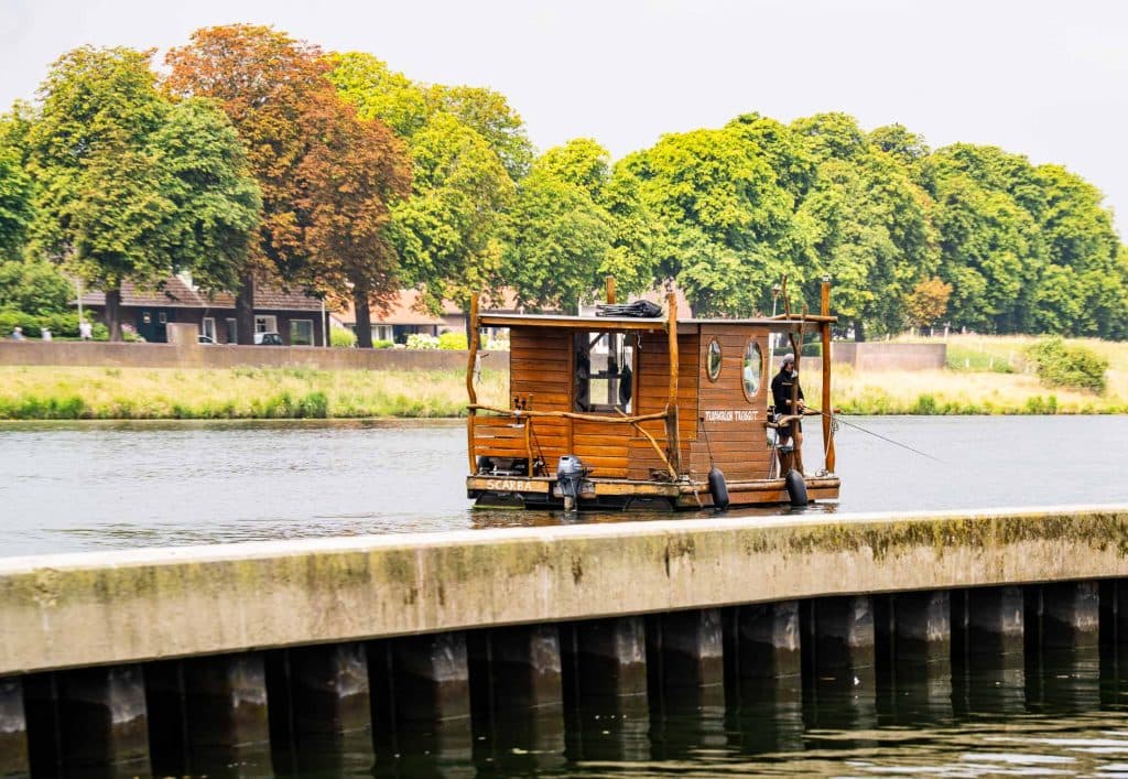 Een kleine houten woonboot met hengels ligt aangemeerd aan een rustige rivier, waarbij traditionele charme naadloos samengaat met automatische concepten. Op het dek staat iemand die geniet van de serene sfeer. Op de achtergrond zijn groene en bruine bomen en gebouwen gedeeltelijk zichtbaar achter de oeverwal.