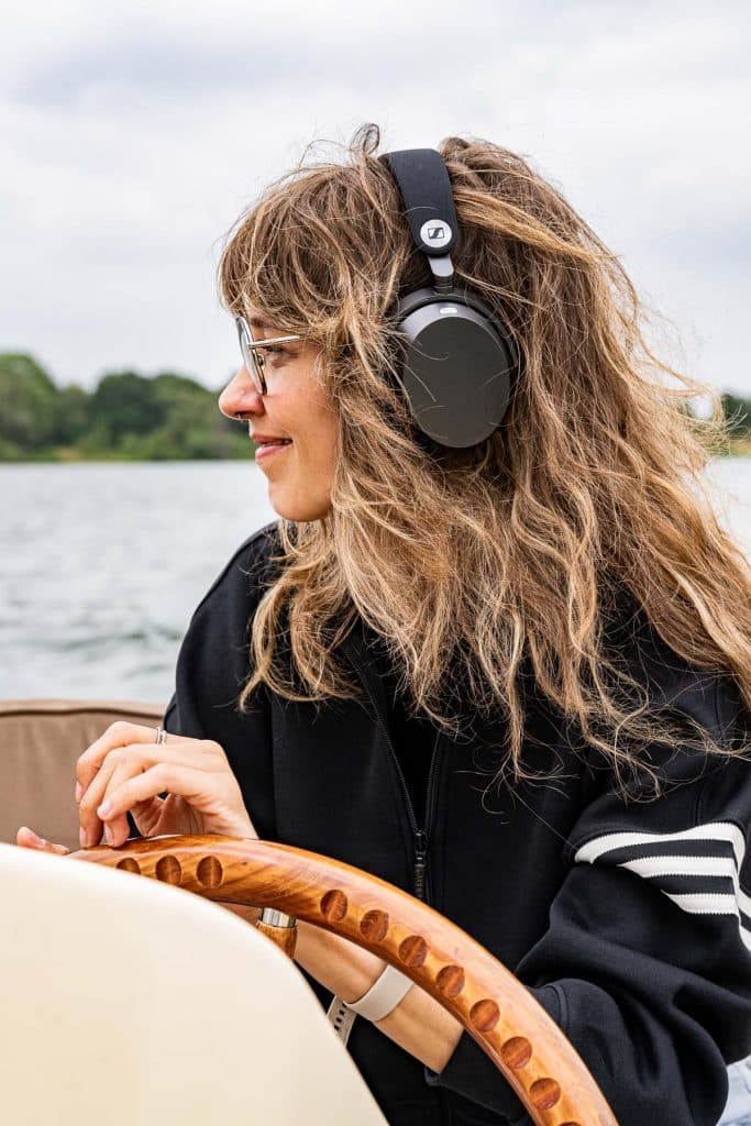 Een vrouw met golvend haar en een bril, met een zwarte koptelefoon op en gekleed in een zwart jasje met witte strepen, zit buiten aan het water; ze glimlacht en houdt het stuurwiel van een houten boot vast – een beeld dat de naadloze stroom van automatische concepten oproept. Op de achtergrond zijn bomen en een bewolkte hemel te zien.