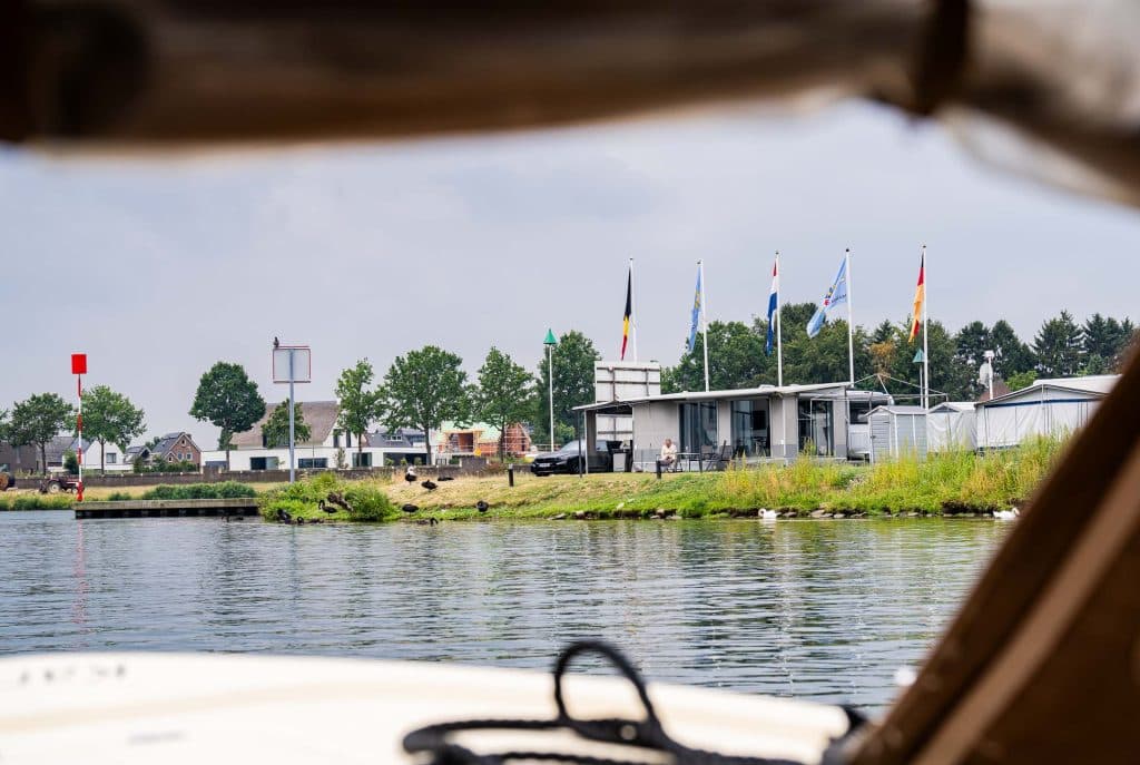 Uitzicht vanaf een boot in de richting van een jachthaven aan de rivier, met op de achtergrond een modern gebouw, diverse vlaggen, een met gras begroeide oever en bomen onder een bewolkte hemel. Het water is kalm en de rand van de boot – die verwijst naar automatische concepten in scheepsontwerp – is op de voorgrond zichtbaar.