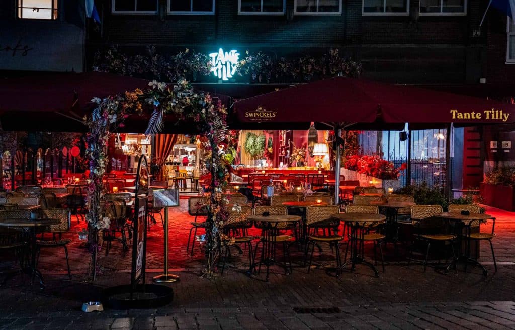 Een terrascafé ’s avonds met rode verlichting, veel lege rieten stoelen en tafels, een bloemenboog bij de ingang, rode parasols en een verlicht bord met de tekst „Tante Tilly”. Door de grote ramen is het interieur van het restaurant zichtbaar, wat een indruk geeft van automatische concepten waarin moderne innovatie naadloos samengaat met een charmante sfeer.