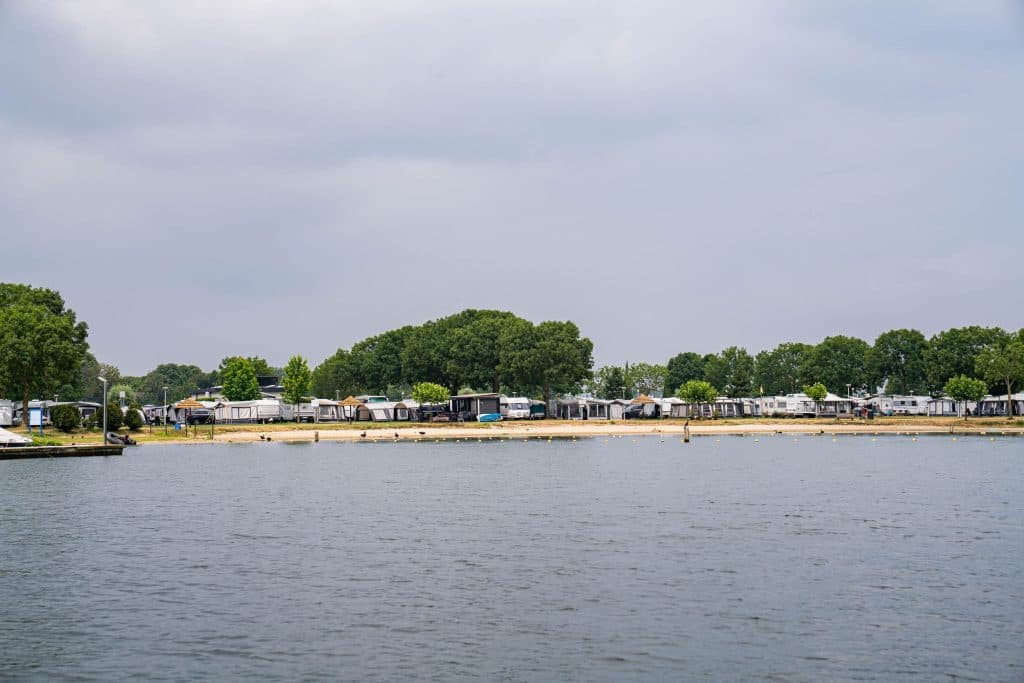 Aan de overkant van het water is een oever te zien met stacaravans en vakantiehuisjes, met op de achtergrond bomen onder een bewolkte hemel – een uitnodigend tafereel voor een ontspannen weekendje Roermond.