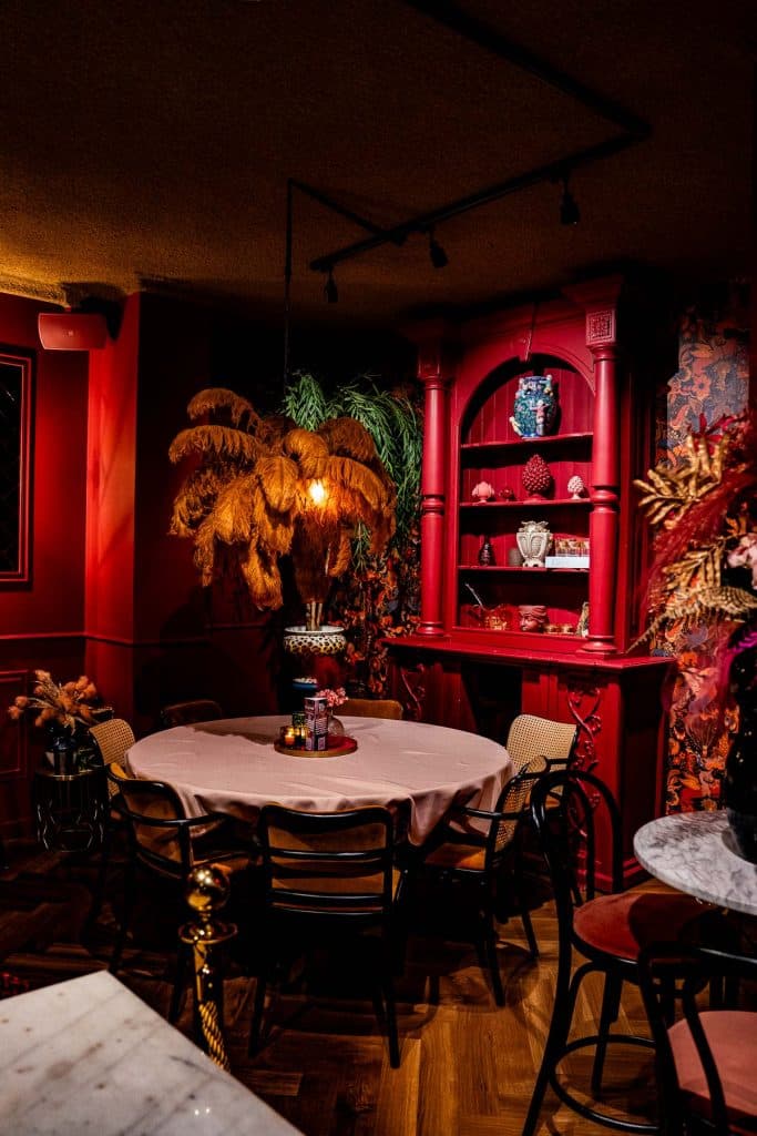 In een schemerige kamer met rode muren staat een ronde tafel met stoelen, een rode vitrinekast met vazen en decoratieve gedroogde planten, wat een gezellige, vintage sfeer creëert die doet denken aan de unieke charme die je zou kunnen ontdekken na een dagje winkelen in Roermond.