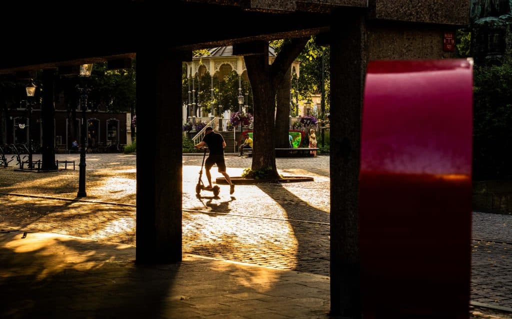 Iemand rijdt op een scooter over een zonovergoten plein met kasseien in het winkelgebied van Roermond, omlijst door donkere pilaren en met op de voorgrond een rood bouwwerk.