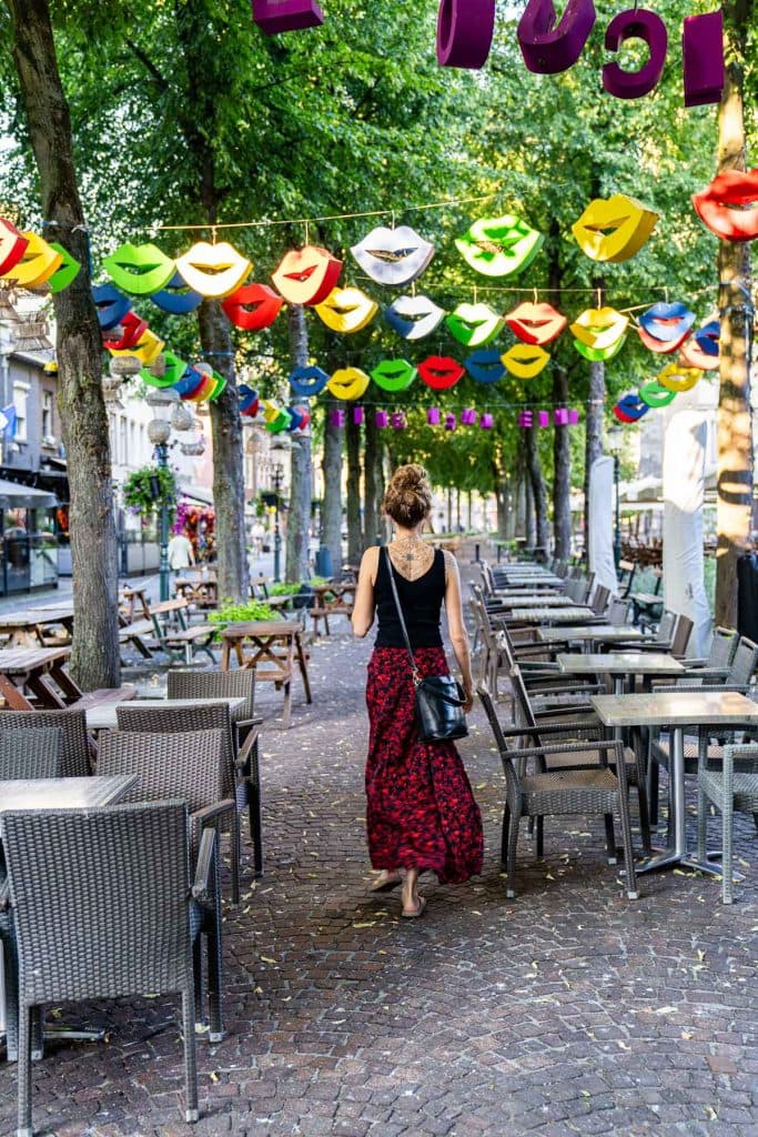 Een vrouw loopt door een terrasgedeelte met lege tafels, onder kleurrijke, lippenvormige versieringen die aan de bomen hangen, en geniet van de levendige sfeer van de nabijgelegen winkelstraat in Roermond.