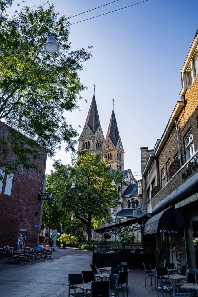 Een rustige straat met terrasjes leidt naar een grote kerk met twee spits torens, omringd door bomen onder een strakblauwe hemel – een perfecte plek om even tot rust te komen na een dagje winkelen in Roermond.