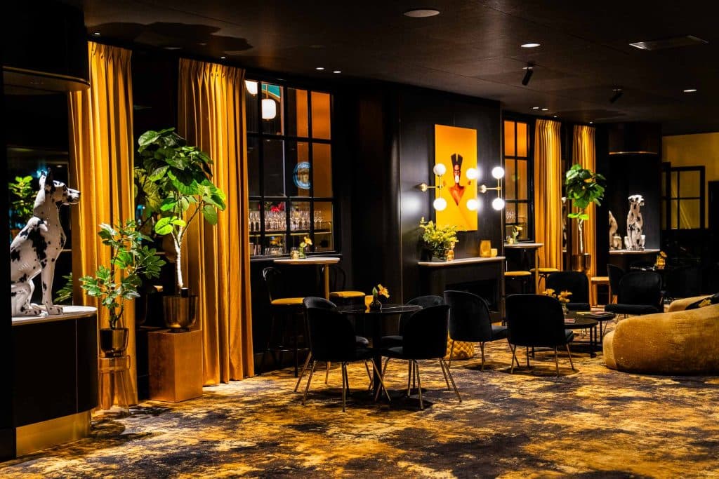 Moderne lounge met gouden gordijnen, zwarte stoelen, ronde tafels, potplanten, dierenbeeldjes en een bar op de achtergrond. Warme verlichting en een tapijt met patroon zorgen voor een gezellige sfeer – perfect om te ontspannen na een dagje winkelen in Roermond.