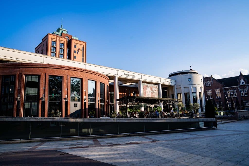 Een modern plein in het winkelgebied van Roermond met bakstenen en afgeronde gebouwen, terrasjes en een strakblauwe lucht op de achtergrond.