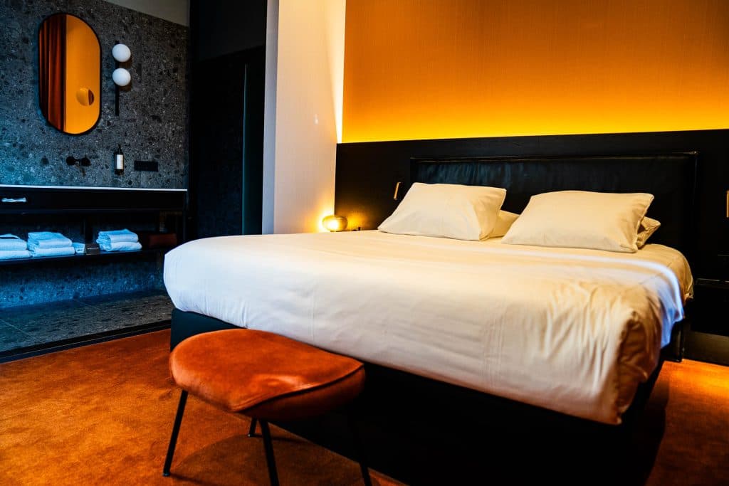 Moderne hotelkamer met een groot bed, wit beddengoed, een oranje accentmuur, een oranje bankje aan het voeteneinde en een badkamermeubel met een ronde spiegel en opgevouwen handdoeken in het zicht – ideaal om te ontspannen na een dagje winkelen in Roermond.