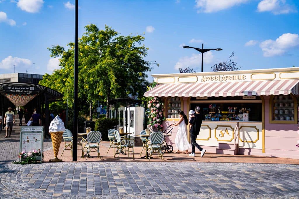 Een dessertkraam in de open lucht met het opschrift „Sweet Avenue”, met lege stoelen en tafels, een grote uitstalling van ijshoorntjes en twee voorbijlopende mensen onder een zonnige, gedeeltelijk bewolkte hemel in het bruisende winkelgebied van Roermond.