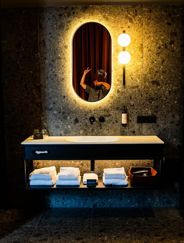 Moderne badkamer met een verlichte ovale spiegel, wandlampen, een wastafel, handdoeken op planken en een persoon die in de spiegel het vredesteken maakt – perfect om te ontspannen na een dagje winkelen in Roermond.