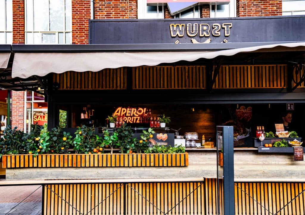 Een eetkraam in de open lucht met de naam „WURST”, voorzien van een baropstelling, een neonreclame met de tekst „APEROL SPRITZ”, decoratieve planten en uitgestalde drankjes in flessen – ideaal om even te pauzeren tijdens je winkeltocht in Roermond.