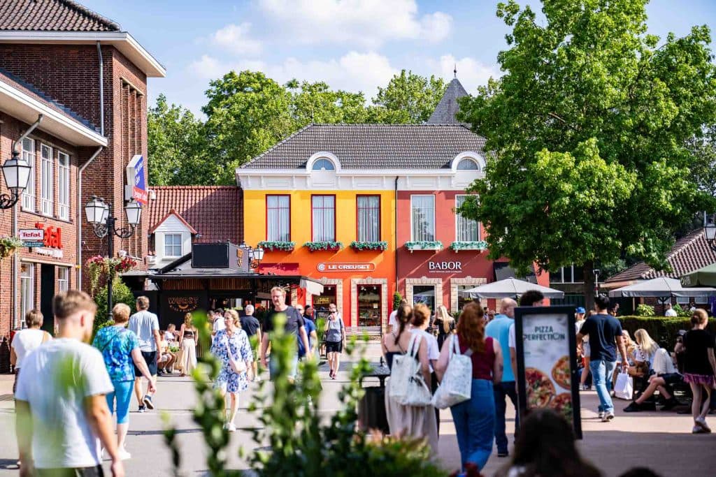 Op een zonnige dag lopen mensen door een levendige straat vol winkels en restaurants, genietend van de bruisende sfeer die zo kenmerkend is voor het winkelen in Roermond, met op de achtergrond een felgeel gebouw en groene bomen eromheen.