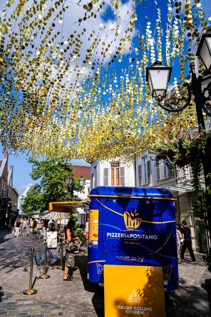 Een blauwe Italiaanse foodtruck staat geparkeerd in een straat met kasseien in het hart van het winkelgebied van Roermond, met mensen in de buurt en gouden versieringen die tussen de gebouwen boven de hoofden hangen.
