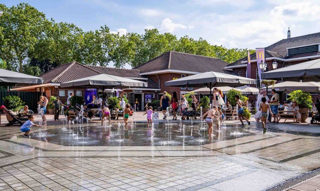 Kinderen spelen bij een fontein op een openbaar plein, omringd door volwassenen die aan tafeltjes onder parasols zitten, met bomen en gebouwen op de achtergrond – een levendig tafereel dat de gezinsvriendelijke sfeer van het winkelgebied van Roermond goed weergeeft.