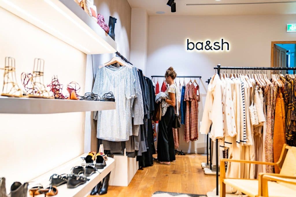 Een vrouw bekijkt de kledingrekken in een goed verlichte boetiek met het opschrift „ba&sh“ aan de muur; op de schappen staan schoenen uitgestald, wat de chique sfeer van het winkelen in Roermond goed weergeeft.