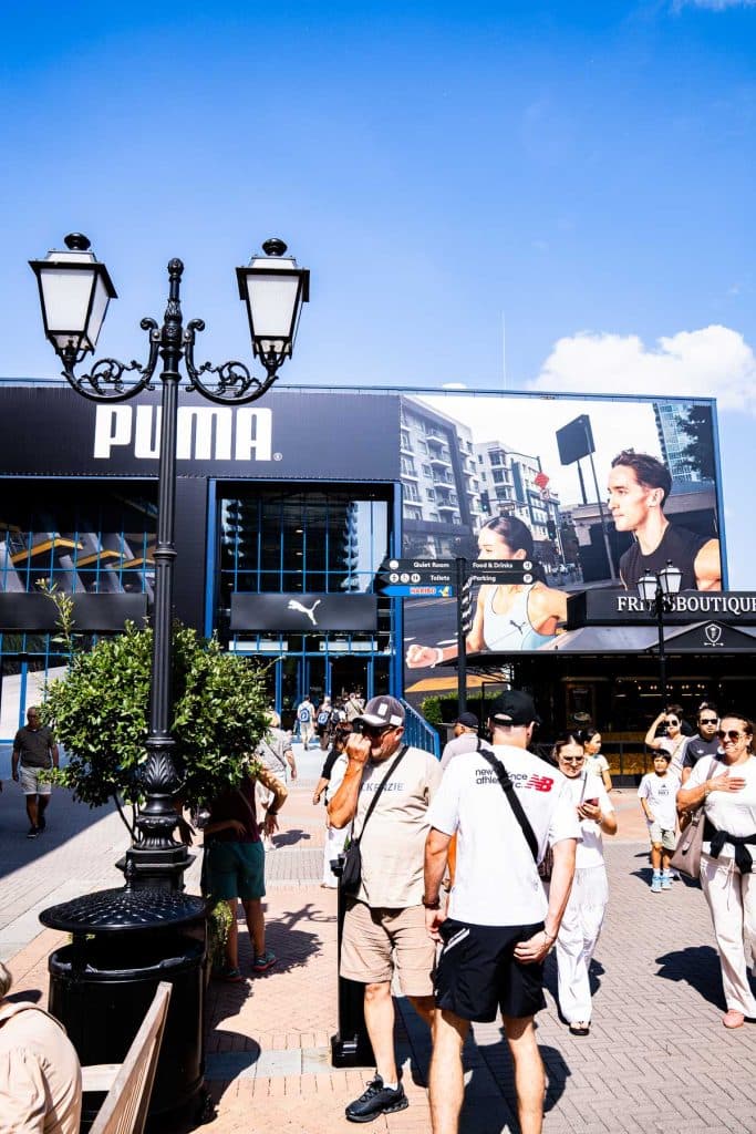 Voor een Puma-winkel in Roermond Shopping zijn mensen te zien die genieten van een zonnige dag, met op de achtergrond een groot reclamebord en een straatlantaarn.