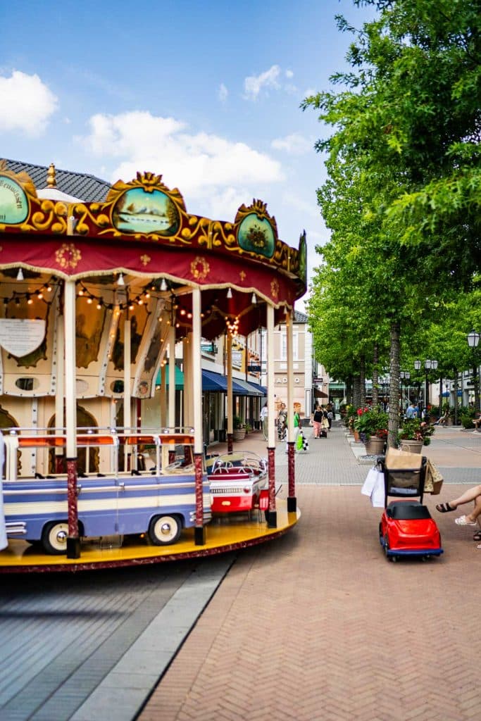In een voetgangerszone van het winkelgebied van Roermond staat een draaimolen met autovormige zitjes. Vlakbij duwt iemand een kind in een rood wagentje, terwijl bomen en winkels de straat omzomen onder een blauwe hemel.