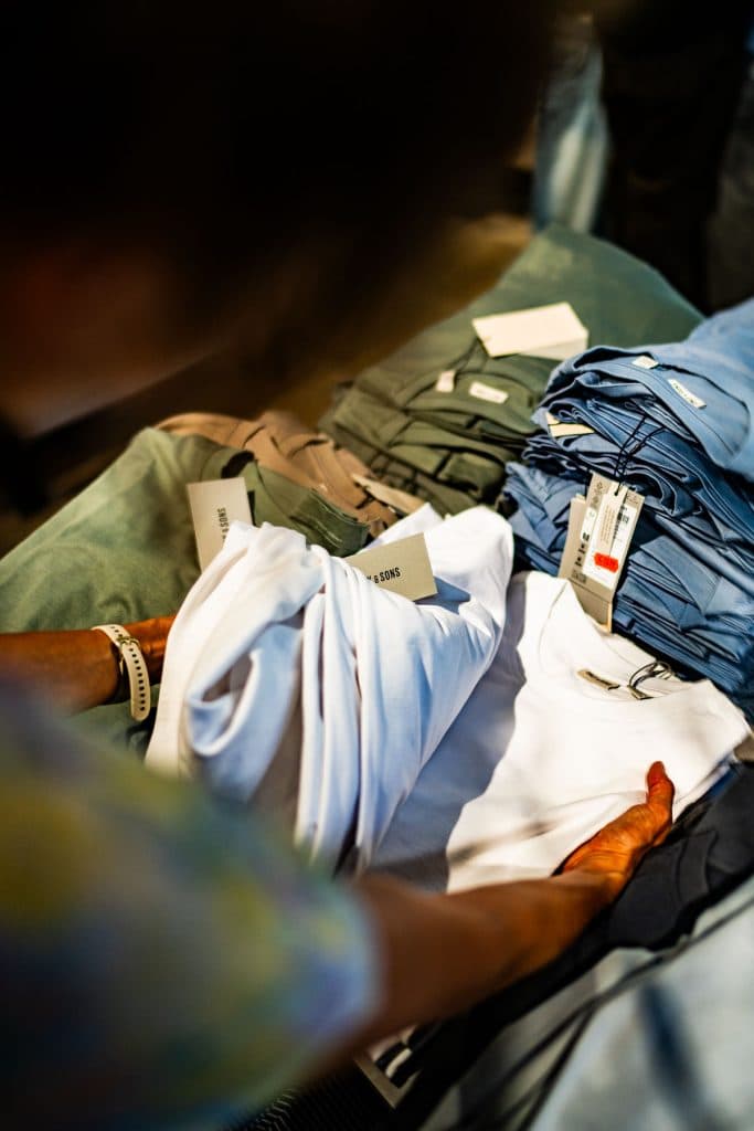 Iemand houdt een opgevouwen wit T-shirt boven een tafel met stapels opgevouwen shirts in verschillende kleuren, elk met zichtbare labels — een beeld dat de overzichtelijke en stijlvolle sfeer van het winkelen in Roermond perfect weergeeft.