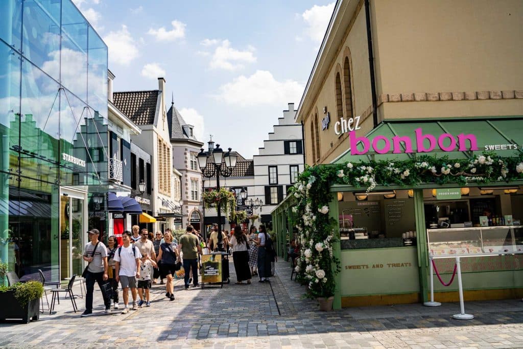 Mensen slenteren over een geplaveide straat met aan weerszijden winkels, waaronder een groene snoepkraam met het bordje „chez bonbon”, en genieten van een zonnige dag in het hart van het winkelgebied van Roermond.