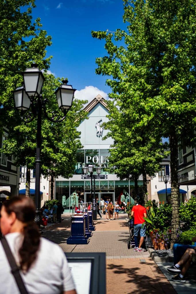 Mensen wandelen en winkelen voor een Polo Ralph Lauren-winkel, omringd door bomen en bankjes, en genieten op een zonnige dag van de levendige winkelsfeer in Roermond.