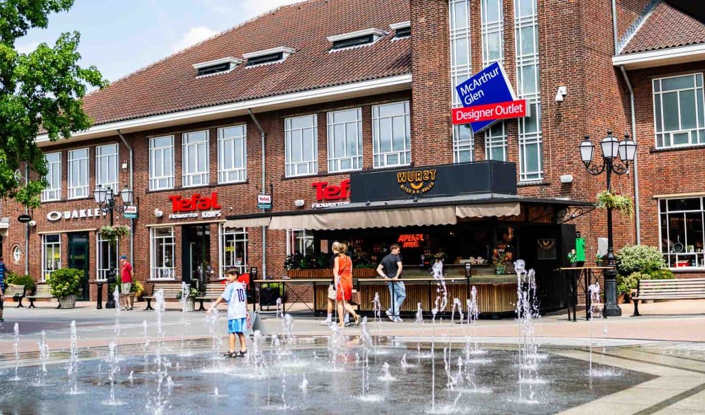 Op een zonnige dag spelen kinderen in een fontein bij een eetkraampje voor de bakstenen outletwinkels, waaronder Oakley en Tefal, in het MacArthur Glen Designer Outlet in Roermond – een populaire bestemming om te winkelen in Roermond.