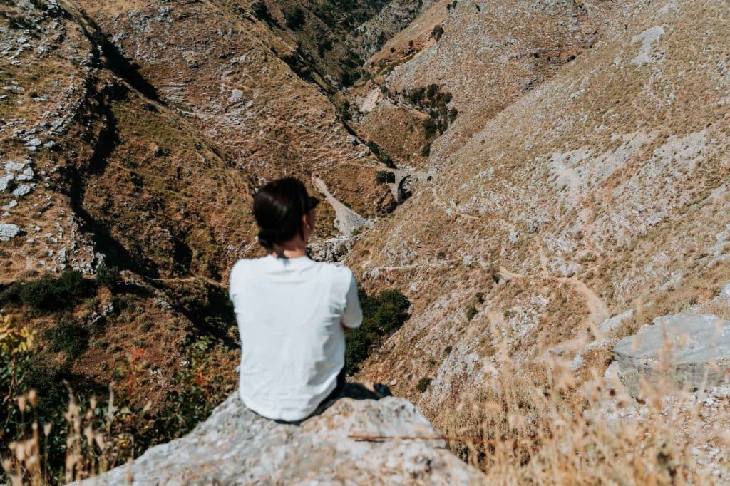 Een persoon met donker haar in een wit overhemd zit op een rots en kijkt uit over een dor, bergachtig landschap met kronkelende paden beneden – een beeld dat de geest van de Albanese nomaden en de vrijheid die je tijdens elke roadtrip door Albanië ervaart, perfect weergeeft.