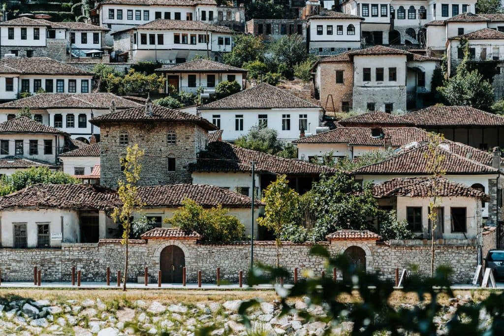 Rijen witte historische huizen met rode pannendaken, gebouwd op een heuvel, omgeven door groen en stenen muren – een schilderachtig tafereel dat erop wacht om ontdekt te worden tijdens je volgende roadtrip door Albanië.
