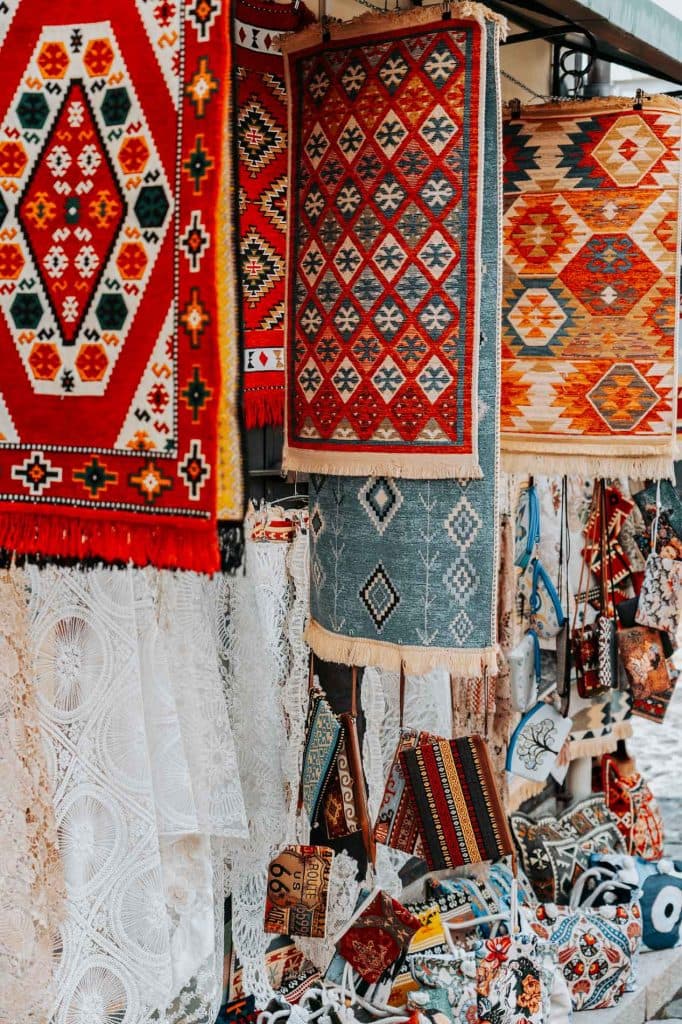 Op een openluchtmarkt worden kleurrijke tapijten met patronen en textiel met geometrische motieven tentoongesteld, naast geborduurde tassen en kanten stoffen – wat een kijkje biedt in de rijke tradities van de Albanese nomaden voor wie aan een roadtrip door Albanië begint.