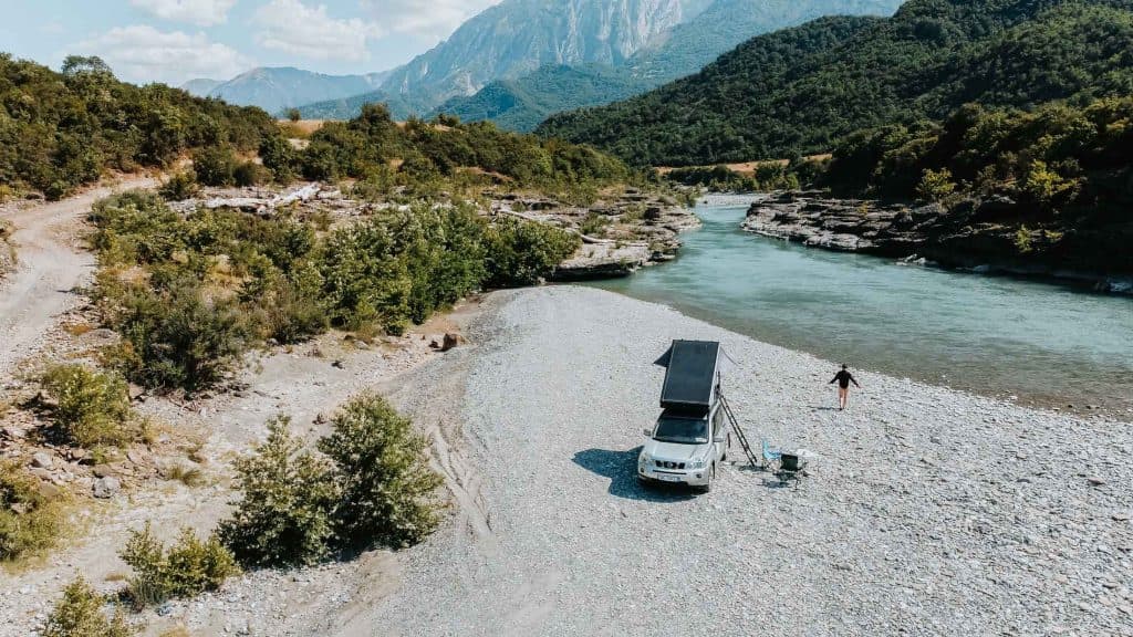 Een witte SUV met een daktent staat geparkeerd op een rotsachtige rivieroever, omringd door groene heuvels en bergen onder een gedeeltelijk bewolkte hemel – een idyllische tussenstop voor alle Albanië-nomaden die tijdens hun roadtrip door Albanië de wilde schoonheid van de regio verkennen.