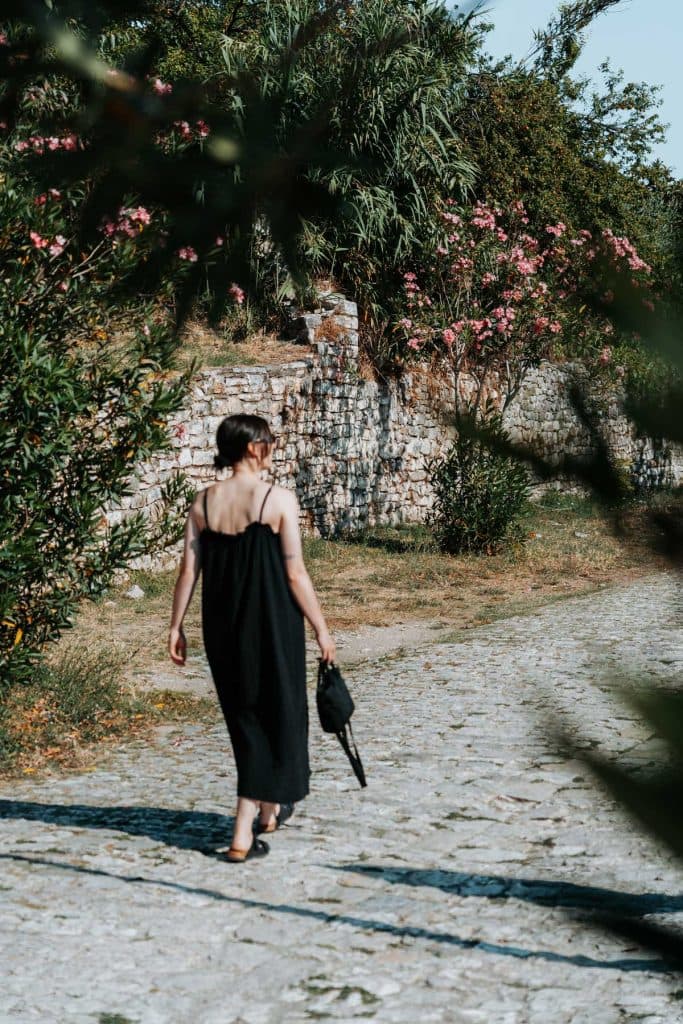 Op een zonnige dag loopt iemand in een zwarte jurk over een stenen pad dat wordt omzoomd door struiken en een bakstenen muur met roze bloemen, waarmee de sfeer wordt weergegeven van de Albanese nomaden tijdens hun roadtrip door Albanië.