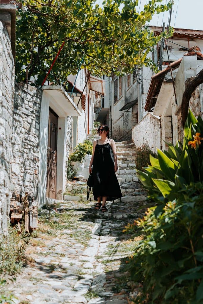 Een persoon in een zwarte jurk loopt over een smal stenen pad, omzoomd door planten en oude stenen gebouwen, in een zonnig Albanees dorpje, en legt daarmee de sfeer vast van een roadtrip door Albanië en de rustige charme waar Albanees reizigers vaak naar op zoek zijn.