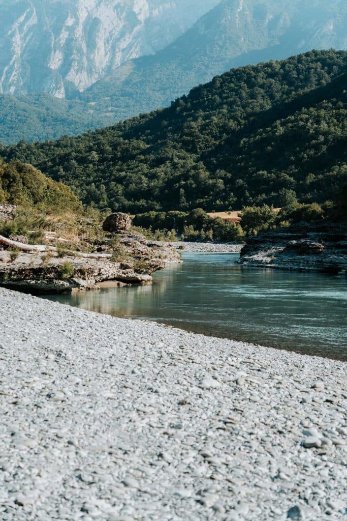 Een heldere rivier stroomt tussen rotsachtige oevers en beboste heuvels, met op de achtergrond bergen die zich aftekenen tegen een blauwe hemel – een uitnodigend tafereel voor elke roadtrip door Albanië, dat de geest weergeeft van de Albanese nomaden die op zoek zijn naar avontuur te midden van de schoonheid van de natuur.
