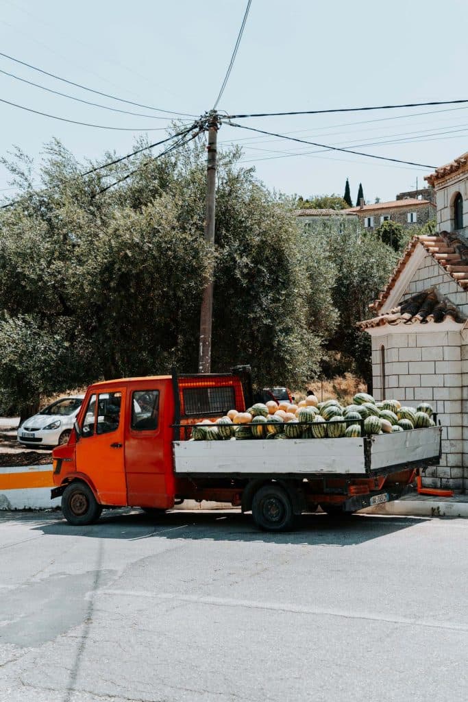 Op een zonnige dag staat er een rode vrachtwagen volgeladen met watermeloenen en andere meloenen langs de kant van de weg, vlakbij bomen en gebouwen, wat de levendige sfeer oproept van een roadtrip door Albanië met Albanese nomaden.
